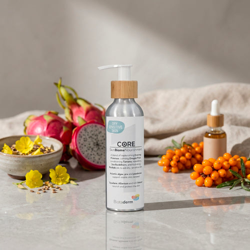 A bottle of Biotaderm Core Dry Sensitive Everyday Moisturiser on a stone surface with dragon fruit, sea buckthorn, yellow flowers, and a dropper bottle in the background—specially formulated with nourishing plant oils to deeply hydrate dry sensitive skin, making it an ideal body moisturiser.