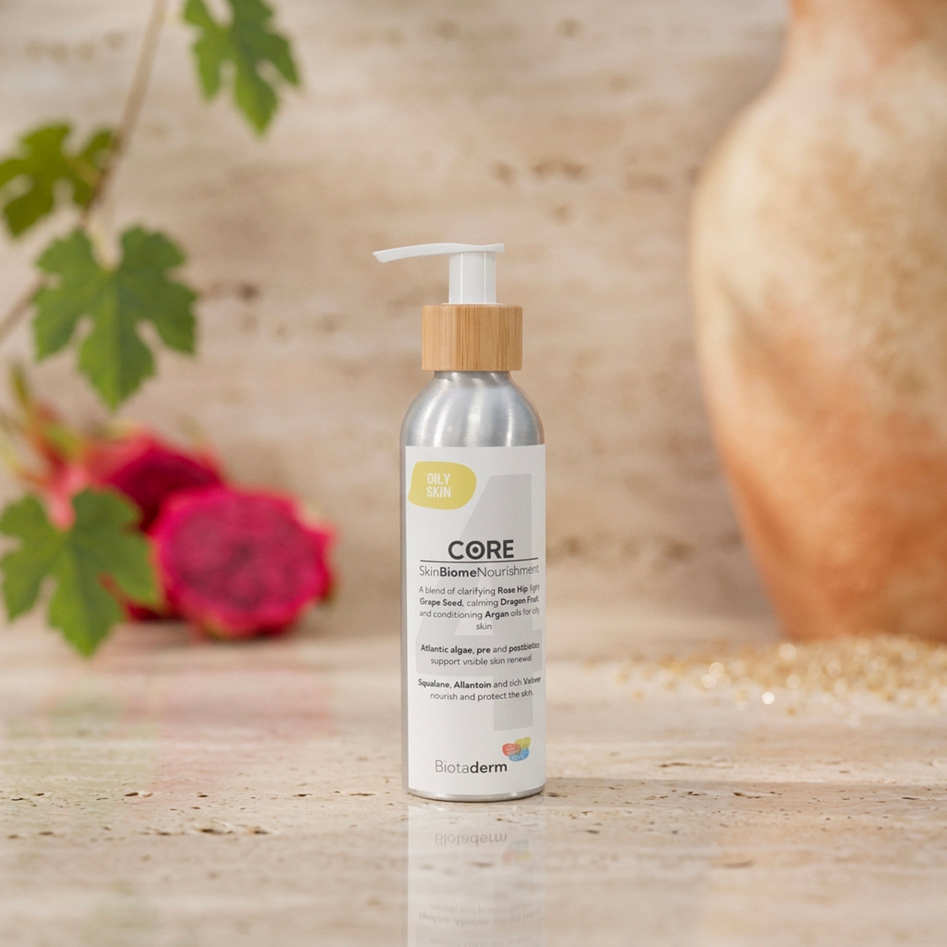 A silver bottle of Biotaderm Core Oily Everyday Moisturiser with a pump sits on a marble surface, enriched with nourishing plant oils—ideal even for oily skin. A blurred flower and vase provide a soft, elegant backdrop.