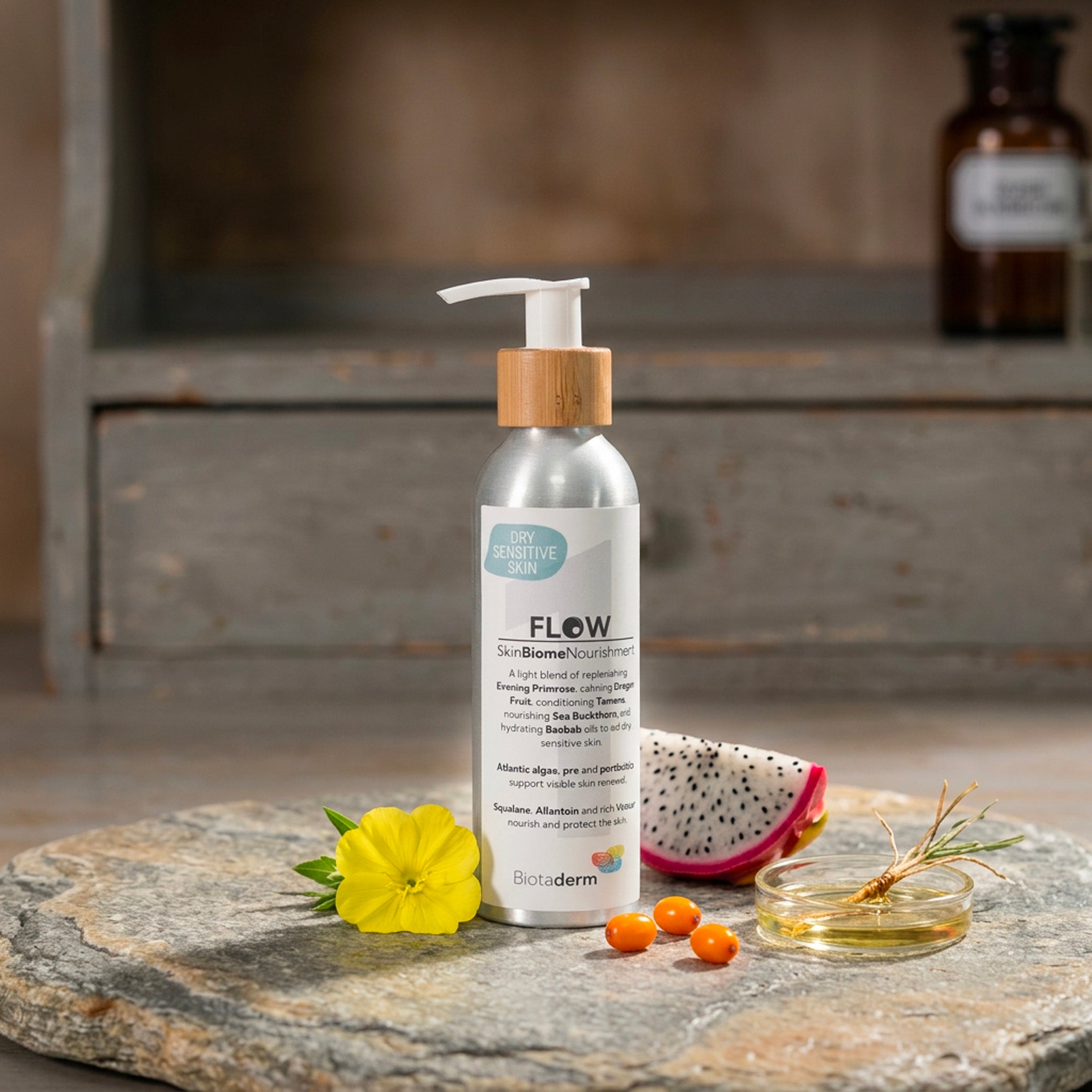 A bottle of Biotaderm Flow Dry Sensitive Light Moisturiser rests on a stone surface alongside dragon fruit, yellow flowers, orange capsules, and a small dish of oil—showcasing this body moisturiser’s unique blend of plant oils formulated to deeply nourish dry sensitive skin.