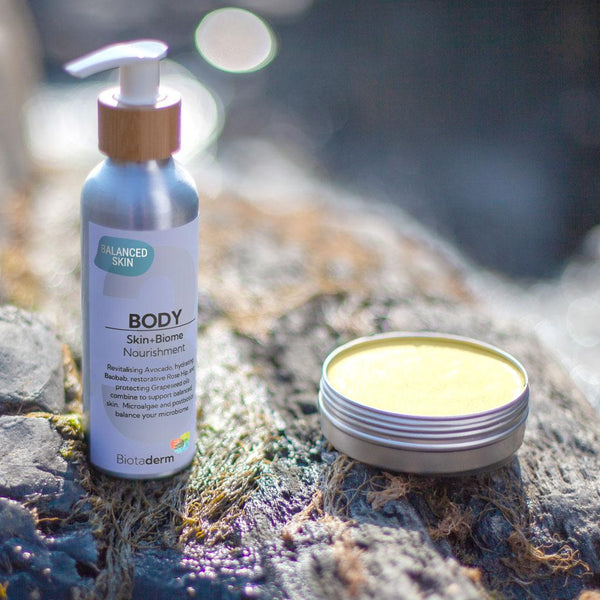 A bottle of Biotaderm's Body Moisturiser - Balanced and an open tin of cream, enriched with Dragon Fruit Oils, are placed on a rocky surface, illuminated by natural sunlight.